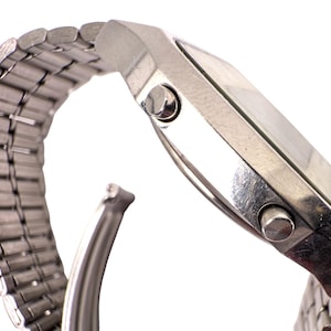 Puede incluir: Un reloj plateado con una esfera rectangular y una correa de metal con eslabones. El reloj tiene dos botones en el lateral y una fijaci&oacute;n de correa curvada. El reloj se muestra sobre un fondo blanco.