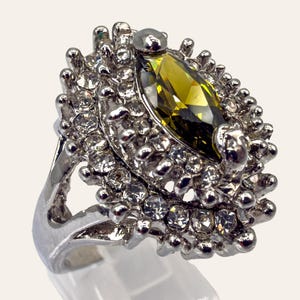 May include: Silver-toned ring featuring a large, olive-green marquise-cut gemstone at the center, surrounded by numerous smaller clear crystals. The ring has a detailed, textured setting with a wide band.