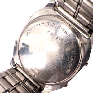 Puede incluir: Primer plano de la parte trasera de un reloj de acero inoxidable con una correa de metal plateado. La esfera del reloj tiene el texto "WATER RESISTANT STAINLESS STEEL A311292 A914-5A09 AO". La correa est&aacute; hecha de peque&ntilde;os eslabones met&aacute;licos rectangulares.