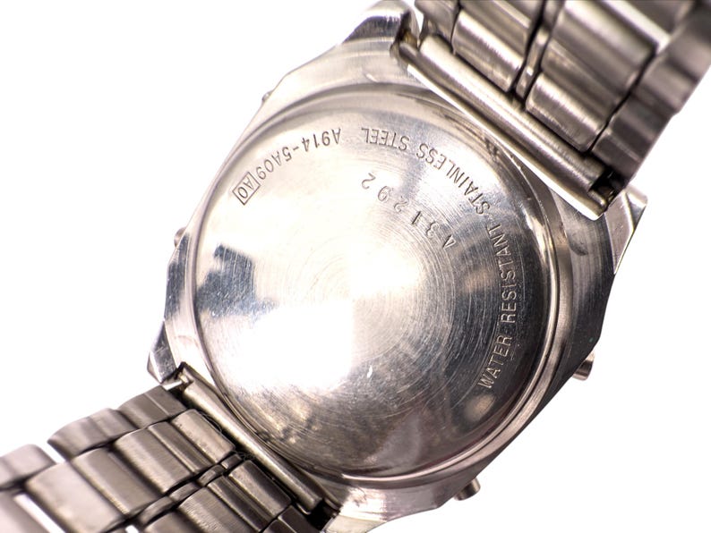 Puede incluir: Primer plano de la parte trasera de un reloj de acero inoxidable con una correa de metal plateado. La esfera del reloj tiene el texto "WATER RESISTANT STAINLESS STEEL A311292 A914-5A09 AO". La correa est&aacute; hecha de peque&ntilde;os eslabones met&aacute;licos rectangulares.