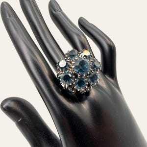 May include: A cluster ring featuring a large central dark blue faceted stone surrounded by a ring of smaller matching blue stones set in a silver-toned metal frame. The stones are arranged in a circular floral pattern with prong settings.