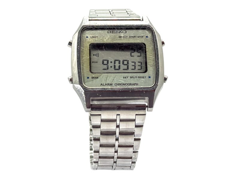 Puede incluir: Un reloj digital Seiko vintage con caja y correa plateadas. La esfera muestra la hora, la fecha y otras funciones. El reloj tiene botones para luz, modo e inicio/parada. Es un cron&oacute;grafo de alarma.