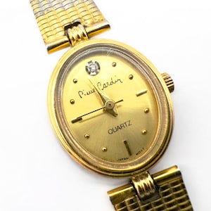 May include: A gold-toned Pierre Cardin quartz watch with an oval face. The watch features a gold band and a gold dial with the word "QUARTZ" and the brand name. The watch has a small diamond-like accent at the 12 o'clock position.