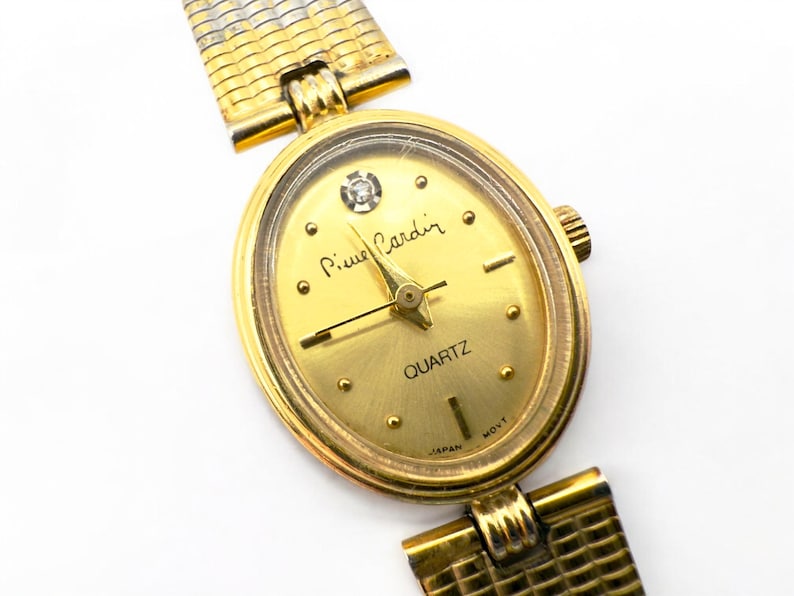 May include: A gold-toned Pierre Cardin quartz watch with an oval face. The watch features a gold band and a gold dial with the word "QUARTZ" and the brand name. The watch has a small diamond-like accent at the 12 o'clock position.