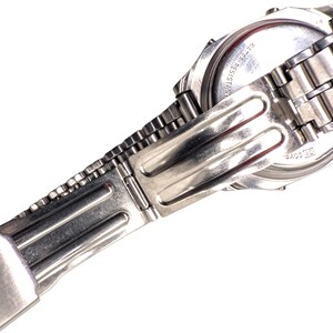 Puede incluir: Primer plano de un reloj de pulsera plateado con una correa de metal. La esfera del reloj es visible, con texto que indica resistencia al agua. La correa est&aacute; conectada con un cierre plegable. El reloj est&aacute; sobre un fondo blanco.