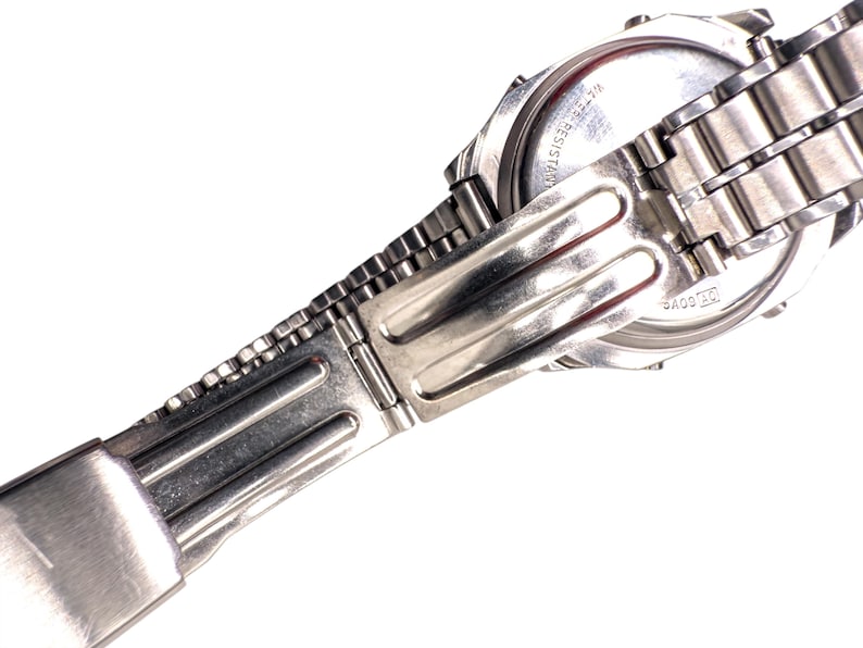 Puede incluir: Primer plano de un reloj de pulsera plateado con una correa de metal. La esfera del reloj es visible, con texto que indica resistencia al agua. La correa est&aacute; conectada con un cierre plegable. El reloj est&aacute; sobre un fondo blanco.