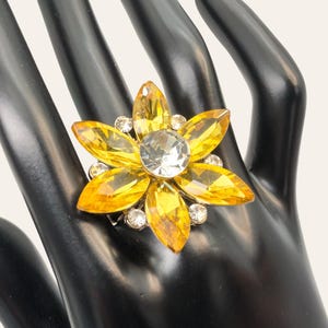 May include: A statement ring featuring a large, flower-shaped design with six marquise-cut yellow crystals surrounding a central round clear crystal. Small, round, clear accent stones are set between the yellow petals on a metallic base.