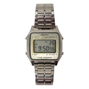 Puede incluir: Un reloj digital Seiko plateado con una esfera rectangular y una correa de metal a juego. El reloj muestra la hora como 9:50:55, con el d&iacute;a y la fecha como Vi 25. El reloj tiene botones para luz, modo y cron&oacute;grafo de alarma.