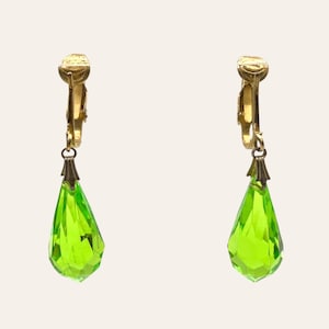 Pode incluir: Um par de brincos de pressão em tom dourado com gotas de cristal verde-limão facetadas em forma de pera. Cada brinco possui um mecanismo de pressão metálico dourado no topo, ligado ao pendente de cristal por uma pequena tampa metálica.