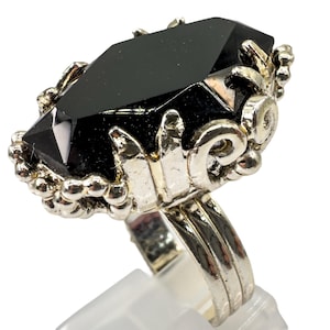 May include: A silver ring with a large, black, faceted gemstone. The gemstone is rectangular with angled edges. The ring has an ornate, detailed setting with a textured design around the stone.