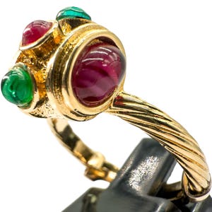 Puede incluir: Un anillo dorado con una gema roja oscura grande y redonda en el centro, flanqueada por dos gemas verdes más pequeñas y dos gemas rojas más pequeñas. La banda tiene un diseño de cuerda trenzada.