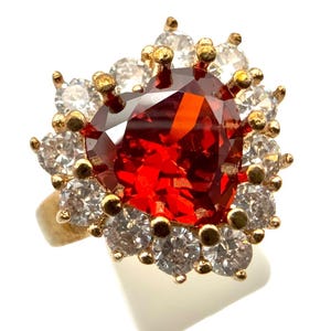 May include: A gold ring with a large, heart-shaped, red gemstone at the centre, encircled by smaller, clear gemstones. The ring's band is plain, polished gold.