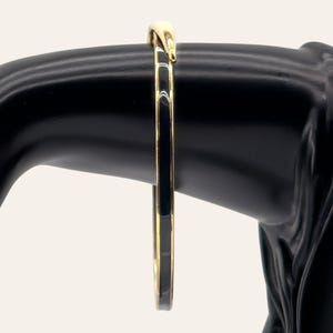 Op de afbeelding: Een dunne, ronde armband met een gepolijst goudkleurig metalen frame en een gladde, glanzende zwarte emaille inleg die langs het midden van de band loopt. Het ontwerp is eenvoudig en strak, met een doorlopende, uniforme metalen rand.