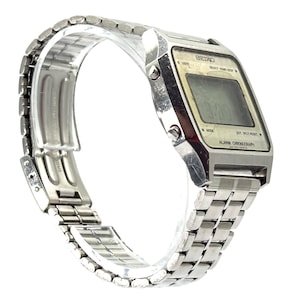 Puede incluir: Un reloj digital Seiko vintage con una correa y caja de metal plateado. La esfera del reloj muestra la hora y tiene botones para varias funciones. La correa del reloj mide aproximadamente 20 cm de largo.