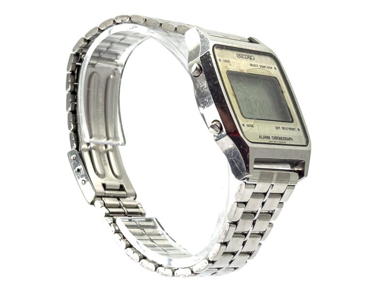 Puede incluir: Un reloj digital Seiko vintage con una correa y caja de metal plateado. La esfera del reloj muestra la hora y tiene botones para varias funciones. La correa del reloj mide aproximadamente 20 cm de largo.
