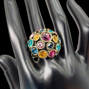 May include: A dome-shaped ring featuring a textured antique gold-tone metal setting embedded with numerous round, multi-coloured faceted crystals in shades of magenta, bright blue, light pink, and amber yellow arranged in a cluster pattern.