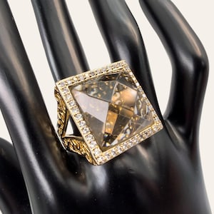 May include: A gold-toned ring featuring a large, square-cut, faceted smoky brown gemstone at the centre. The square setting is bordered by a row of small, clear, sparkling stones. The band displays an intricate, textured, openwork metal design.