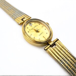 May include: A gold-toned Pierre Cardin quartz watch with a gold oval face and a diamond accent at the 12 o'clock position. The watch has a gold and silver mesh band. The watch face reads "Pierre Cardin" and "QUARTZ".