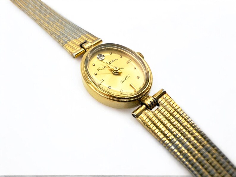 May include: A gold-toned Pierre Cardin quartz watch with a gold oval face and a diamond accent at the 12 o'clock position. The watch has a gold and silver mesh band. The watch face reads "Pierre Cardin" and "QUARTZ".