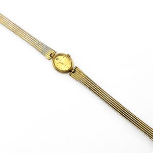 May include: A gold-toned wristwatch with a rectangular face and a gold and silver band. The watch face is a light gold color with small markers. The band is made of thin, flexible metal links. The watch is displayed on a white background.