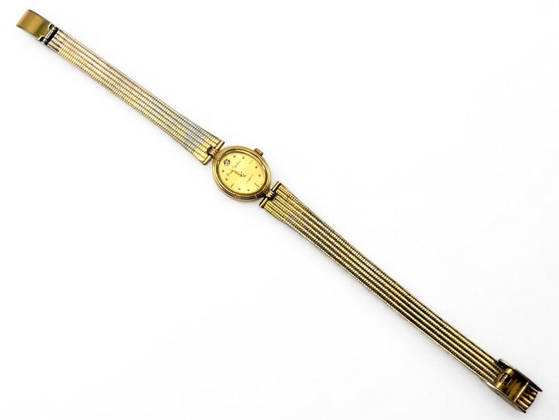 May include: A gold-toned wristwatch with a rectangular face and a gold and silver band. The watch face is a light gold color with small markers. The band is made of thin, flexible metal links. The watch is displayed on a white background.