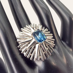 May include: A silver ring with a large, rectangular, light blue gemstone at its centre. The ring has a starburst design with pointed rays extending outwards from the central stone. The ring is displayed on a black hand.