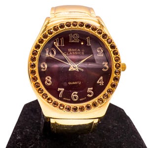 Könnte beinhalten: Goldfarbene Armbanduhr mit dunkelviolettem Zifferblatt und goldenen Ziffern. Die Uhr hat eine Lünette mit kleinen, braunen Edelsteinen. Auf dem Zifferblatt steht "Boca Classics" und "Quartz". Die Uhr liegt auf einer schwarzen Oberfläche.