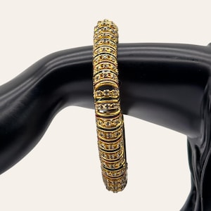 May include: A gold-toned bangle bracelet featuring a series of repeating circular segments. Each segment is embellished with a row of small, clear, sparkling stones set within the metal frame, creating a continuous textured and reflective design.