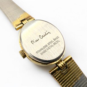 May include: Close-up of a Pierre Cardin wristwatch. The watch has a gold-toned oval face and a gold and silver-toned band. The back of the watch is visible, with the Pierre Cardin signature and text that reads "STAINLESS STEEL BACK BASE METAL BEZEL."