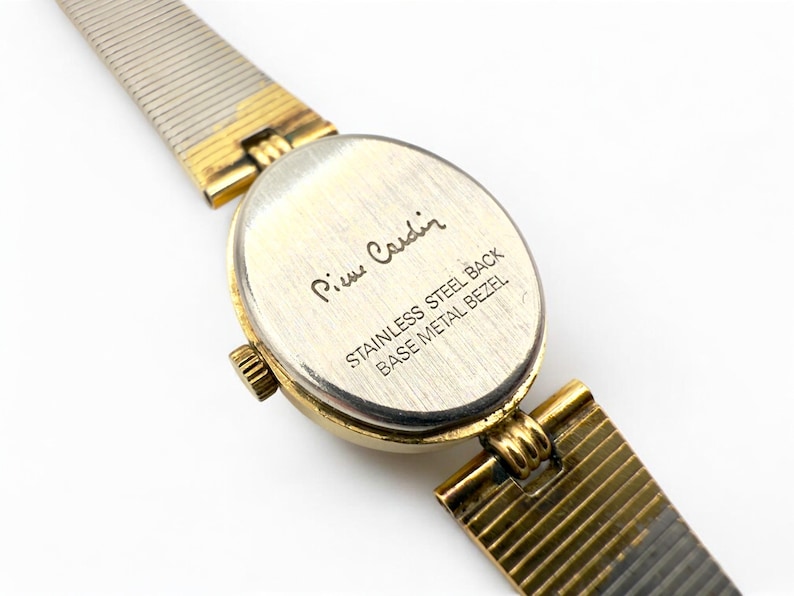 May include: Close-up of a Pierre Cardin wristwatch. The watch has a gold-toned oval face and a gold and silver-toned band. The back of the watch is visible, with the Pierre Cardin signature and text that reads "STAINLESS STEEL BACK BASE METAL BEZEL."