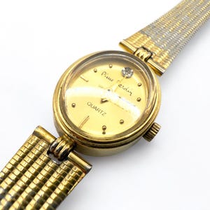 May include: A gold-toned Pierre Cardin quartz watch with a round face and a diamond accent. The watch features a gold-colored dial with gold hour markers and a gold and silver mesh band. The watch is a classic timepiece.