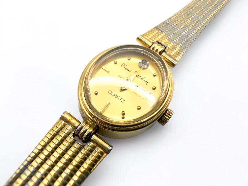 May include: A gold-toned Pierre Cardin quartz watch with a round face and a diamond accent. The watch features a gold-colored dial with gold hour markers and a gold and silver mesh band. The watch is a classic timepiece.