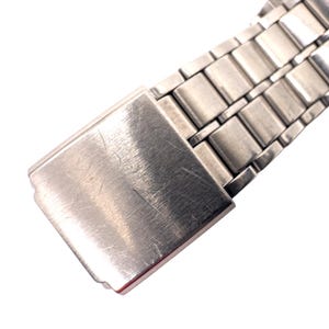 Puede incluir: Primer plano del cierre y los eslabones de una correa de reloj plateada. El cierre es rectangular con un acabado de metal cepillado, que muestra peque&ntilde;os ara&ntilde;azos. Los eslabones de la correa del reloj tambi&eacute;n son plateados y rectangulares, con un aspecto pulido.