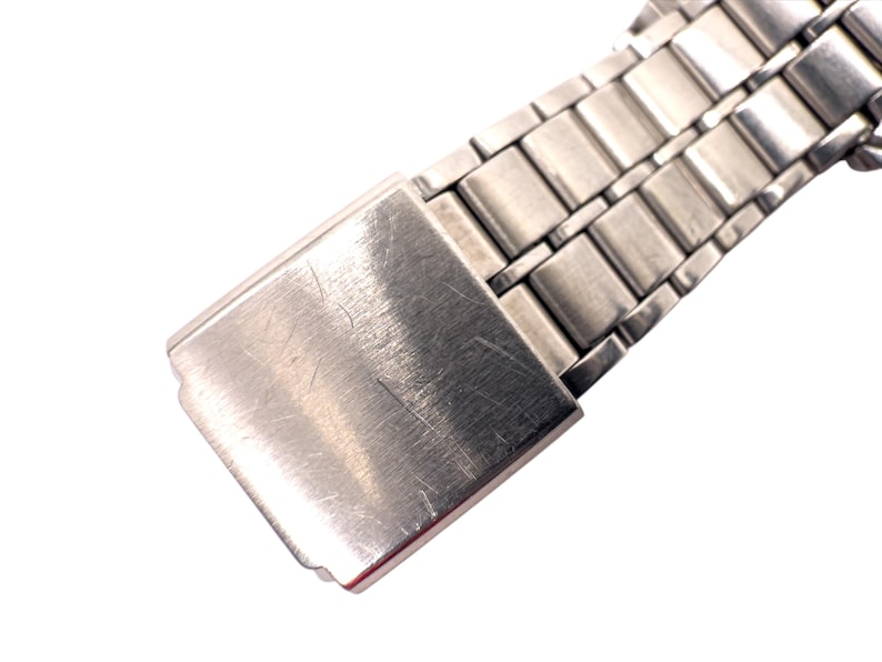 Puede incluir: Primer plano del cierre y los eslabones de una correa de reloj plateada. El cierre es rectangular con un acabado de metal cepillado, que muestra peque&ntilde;os ara&ntilde;azos. Los eslabones de la correa del reloj tambi&eacute;n son plateados y rectangulares, con un aspecto pulido.