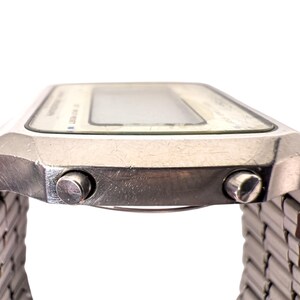 Puede incluir: Primer plano de un reloj digital plateado con una esfera rectangular y una correa de metal. El reloj tiene dos botones laterales y una pantalla clara. Reloj de estilo vintage.
