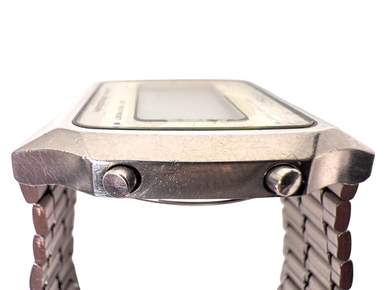 Puede incluir: Primer plano de un reloj digital plateado con una esfera rectangular y una correa de metal. El reloj tiene dos botones laterales y una pantalla clara. Reloj de estilo vintage.