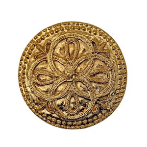 May include: A round, gold-colored button with an intricate design. The button features a central floral-like pattern surrounded by a beaded border. The design is raised, creating a textured surface. The button is likely made of metal.