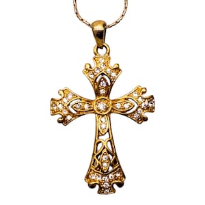 Pode incluir: Um pendente de cruz dourada com um design detalhado, adornado com numerosas pedras preciosas pequenas e transparentes. A cruz é suspensa por uma fina corrente dourada, criando uma joia clássica e elegante.