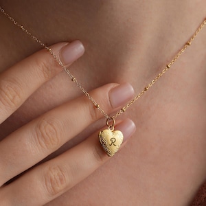May include: A gold heart-shaped locket necklace with a delicate chain and a small initial "R" engraved on the front.
