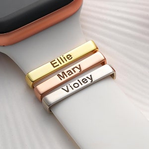 May include: A white Apple Watch band with three gold, rose gold, and silver metal bands with the names "Ellie", "Mary", and "Violey" engraved on them.