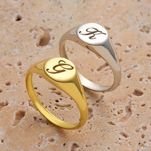 May include: Two signet rings, one gold and one silver, with a cursive letter 'G' engraved on each. The rings are on a textured stone surface.
