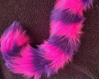 May include: A plush, striped tail in vibrant purple and hot pink. The tail is made of soft, faux fur and has a fluffy, rounded tip. The stripes alternate in color, creating a playful and eye-catching design. This accessory is perfect for cosplay or costume parties.
