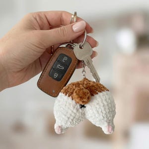 Op de afbeelding: Een novelty sleutelhanger met een gehaakte hondenbil in wit en bruin. De sleutelhanger is bevestigd aan een autosleutel en een gewone sleutel. De hondenbil heeft roze pootjes en een klein zwart kruis.