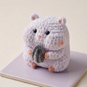 Puede incluir: Un hámster de ganchillo hecho a mano con hilo lavanda suave, sosteniendo una semilla de girasol gris. El hámster amigurumi tiene ojos de seguridad negros, acentos rosas en las orejas, mejillas y pies, y está sobre un cuadrado lavanda. Un regalo hecho a mano lindo y único.