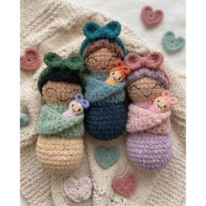 May include: Crocheted dolls in shades of green, blue, and pink, each holding a smaller doll. The dolls have smiling faces and are adorned with bows. Scattered around are crocheted heart shapes in various pastel colors. The dolls rest on a cream-colored knitted blanket.
