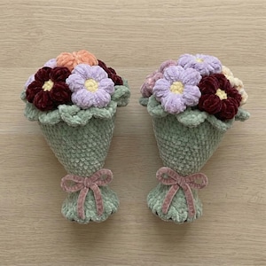 May include: Two handmade crochet flower bouquets. Each bouquet features a variety of colorful flowers in shades of purple, burgundy, peach, and cream, set in a green cone-shaped holder. A pink velvet bow adorns the base of each bouquet.