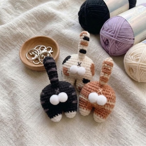 Op de afbeelding: Drie gehaakte kattenkont sleutelhangers in zwart, calico en oranje, elk met een gestreepte staart en pluizige witte pompons. Een kleine houten kom bevat zilveren sleutelringen. Garen in zwart, paars en crème staan op de achtergrond.