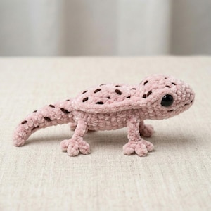 Puede incluir: Un lagarto rosa tejido a mano con ganchillo y manchas marrones oscuras. El reptil de peluche tiene una cabeza redondeada, patas pequeñas y una cola larga. Los ojos del lagarto son negros y está sobre una superficie de color neutro.