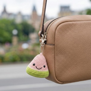 Könnte beinhalten: Ein gehäkelter Wassermelonen-Anhänger in Rosa und Grün mit einem lächelnden Gesicht, der an einer hellbraunen Ledertasche befestigt ist. Der Anhänger ist ein niedliches Accessoire.