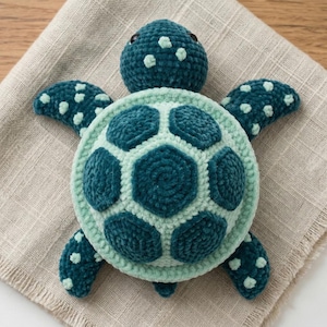 May include: A handmade teal and light green crochet turtle. The turtle has a textured shell with a hexagonal pattern, four flippers, and a small head with two dark eyes. The turtle is lying on a beige cloth.